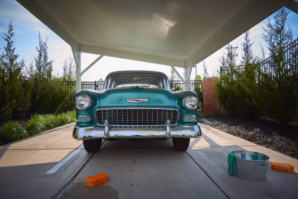 A vintage green Chevrolet car parked under a white carport with bushes and a fence in the background. On the concrete ground in front of the car, there is a metal bucket with a green cloth hanging on it and two orange sponges placed on either side.