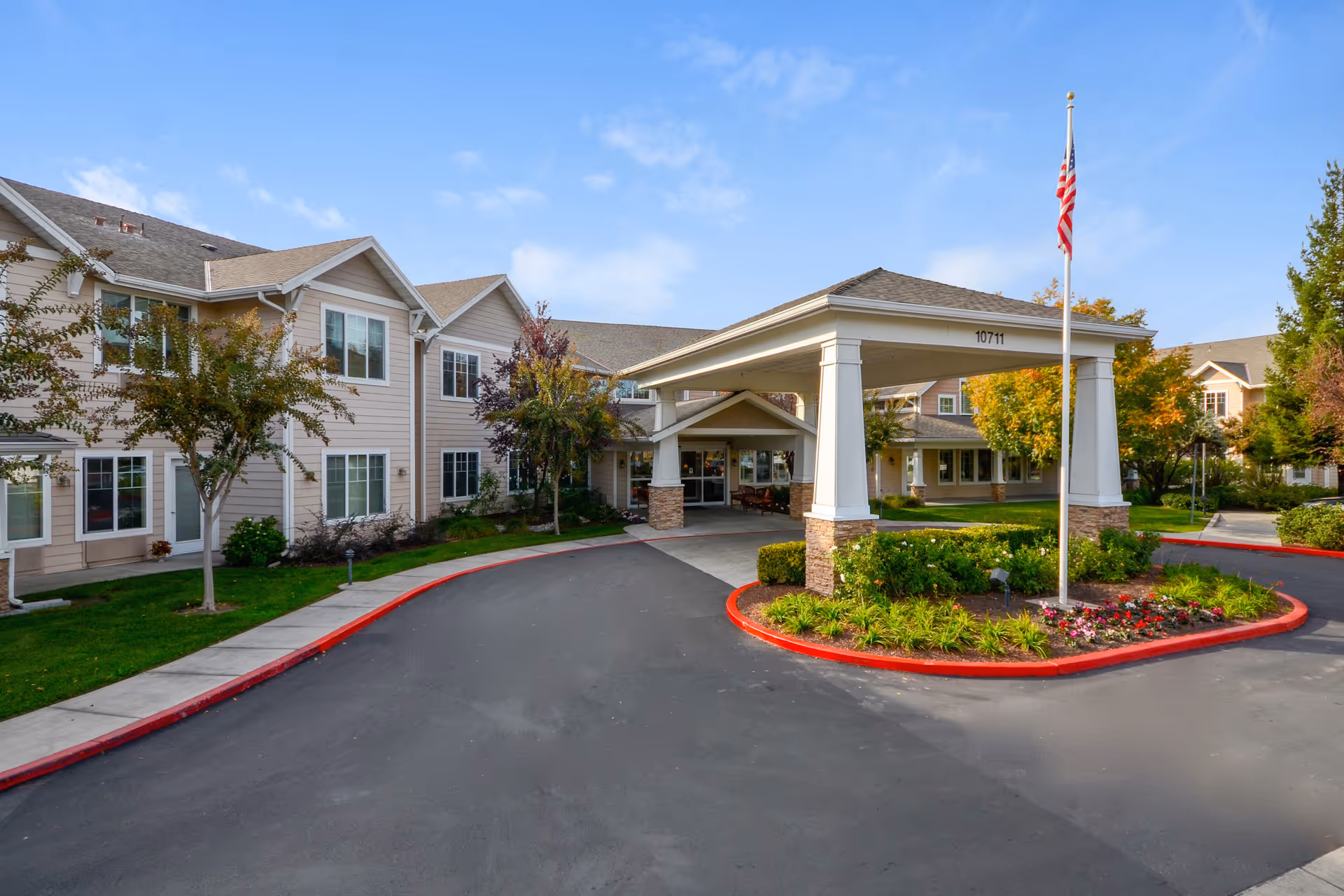 Exterior view of The Commons on Thornton senior living facility showing the main entrance with a covered driveway, an American flag on a flagpole, landscaped flower beds, and a two-story building with multiple windows under a blue sky.