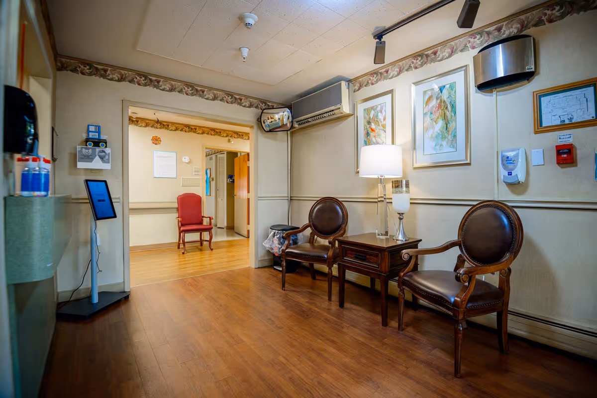 A waiting area inside Pennypack Nursing and Rehabilitation Center featuring two brown leather chairs with wooden frames, a wooden side table with a lamp and decorative candle, and two framed abstract paintings on the wall. The floor is wood, and there is a doorway leading to another room with a red upholstered chair. Various wall-mounted devices and a hand sanitizer dispenser are visible.