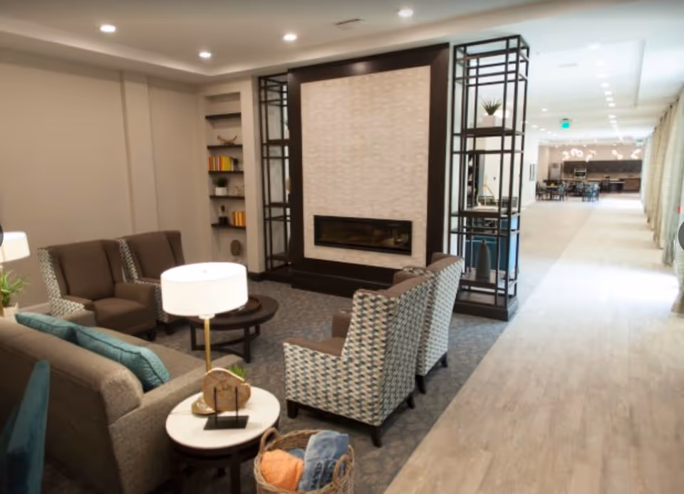 A modern living room area in a senior living facility with comfortable seating including a sofa, armchairs, and a coffee table. There is a contemporary fireplace with a decorative surround, shelving units with plants and books, and a lamp on a side table. The room opens into a bright hallway leading to a dining area and kitchen in the background.