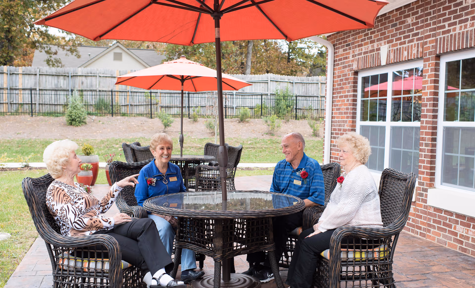 Four elderly people sitting around a round glass-top wicker table under a large red patio umbrella outside a brick building. They are engaged in conversation and appear to be enjoying the outdoor setting with green grass and a wooden fence in the background.