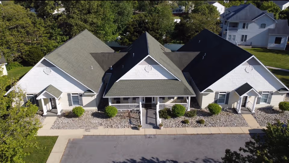 Aerial view of a single-story assisted living facility building with a central entrance porch and two wings on either side. The building has a gray shingled roof, white siding, and small bushes planted along the front. There is a paved driveway and sidewalk leading to the entrances, surrounded by green trees and grass.