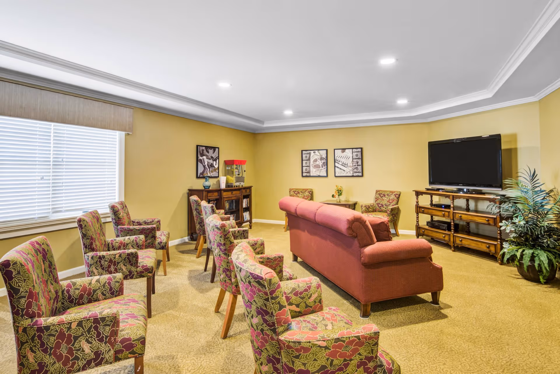 A cozy living room area with a red sofa and multiple floral-patterned armchairs arranged facing a large flat-screen TV on a wooden stand. The room has yellow walls, a window with blinds, framed artwork on the walls, a small table with a flower vase, and a potted plant in the corner.