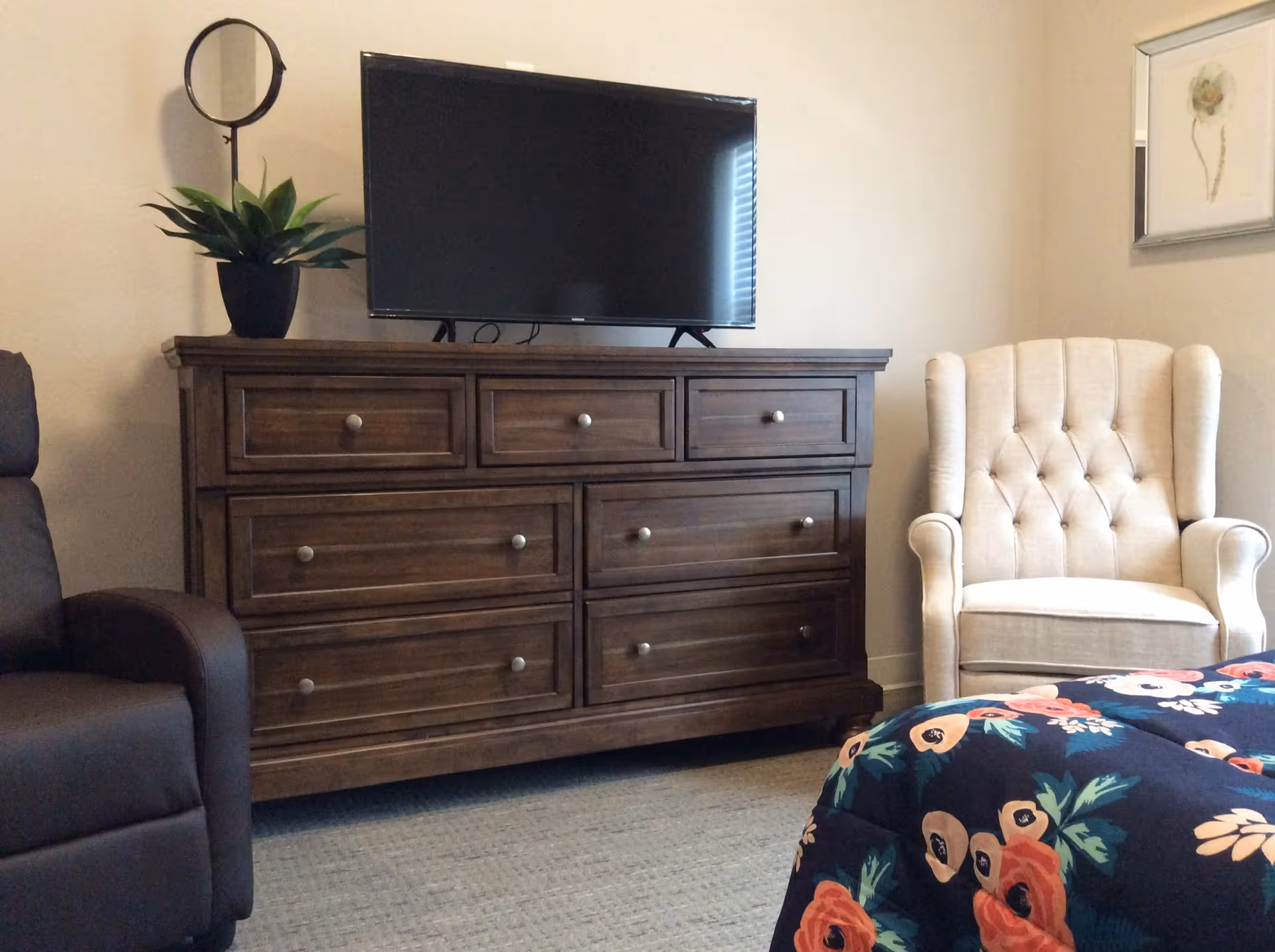 A cozy bedroom corner featuring a dark wooden dresser with a flat-screen TV on top, a potted plant, a beige tufted armchair, part of a floral-patterned bedspread, and a framed flower artwork on the wall.