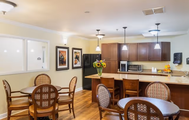 A well-lit kitchen and dining area in an assisted living facility. The room features wooden floors, round tables with patterned cushioned chairs, a kitchen island with three wooden stools, and dark wood cabinets. On the countertop, there is a vase with sunflowers, a microwave, a coffee maker, and a bowl of oranges. The walls are light yellow with two framed pictures hanging. Ceiling lights and pendant lights illuminate the space.