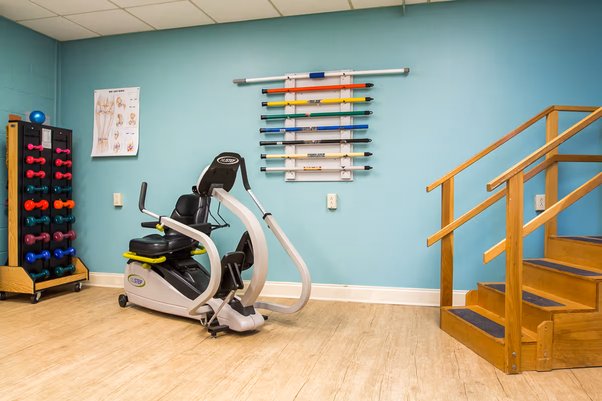 Rehab exercise room with a recumbent stepper machine, a rack of colorful dumbbells, therapy rods mounted on a turquoise wall, and wooden practice stairs.