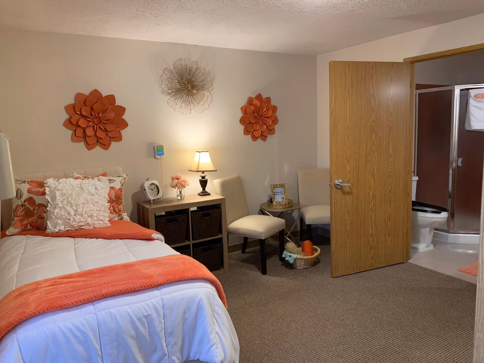 A cozy bedroom in a senior living facility featuring a single bed with white and orange bedding, decorative pillows, and floral wall art above the bed. Next to the bed is a small wooden nightstand with a lamp, clock, and flowers. Two beige chairs and a small round table with a framed photo and basket are positioned near an open door leading to a bathroom with a toilet and shower visible.