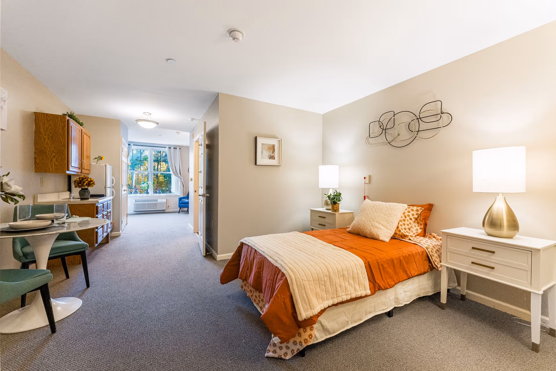 A bright studio apartment interior featuring a made bed with nightstands and lamps, a small kitchenette and dining area, and a windowed sitting area.