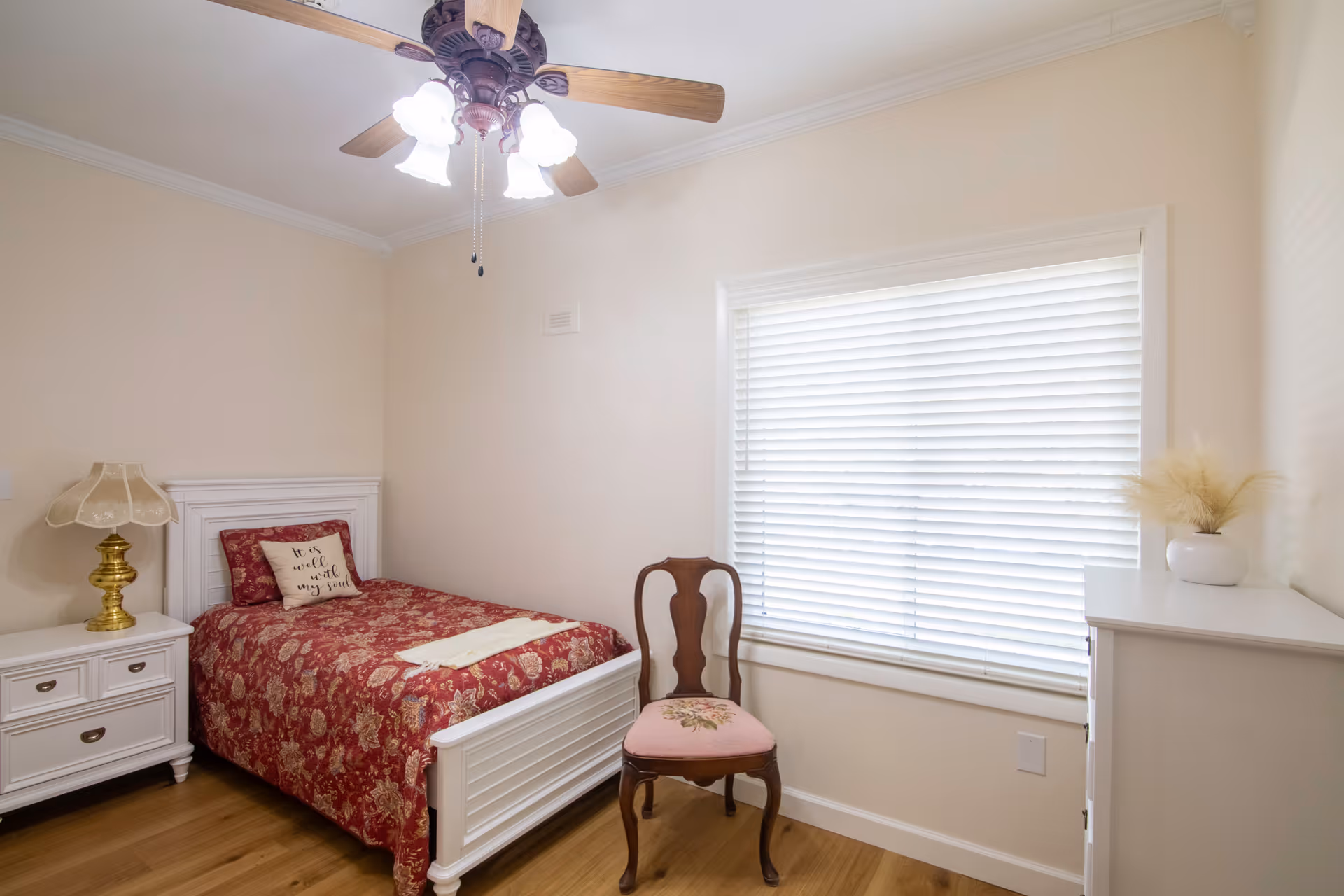 A small bedroom with a single white bed featuring a red floral bedspread and a pillow with the text 'It is well with my soul'. Next to the bed is a white nightstand with a vintage-style lamp. A wooden chair with a pink cushioned seat is placed near a window with white blinds. A white dresser with a small vase of dried flowers sits against the wall. The room has light-colored walls, wooden flooring, and a ceiling fan with lights.