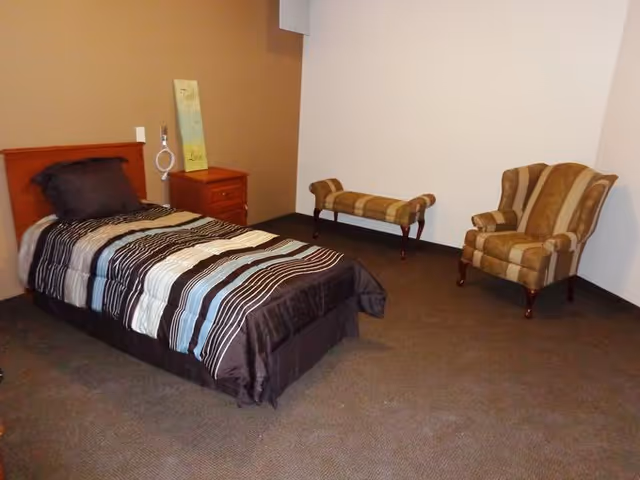 A simple bedroom with a single bed covered in a striped comforter, a wooden nightstand with a decorative sign on it, a striped upholstered armchair, and a matching bench against the wall.