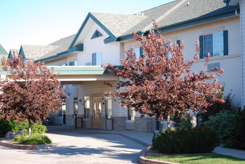 Exterior view of a two-story assisted living facility building with a covered entrance. The building has light-colored siding, multiple windows, and a gray shingled roof. There are two ornamental trees with reddish leaves and some green shrubs near the entrance.