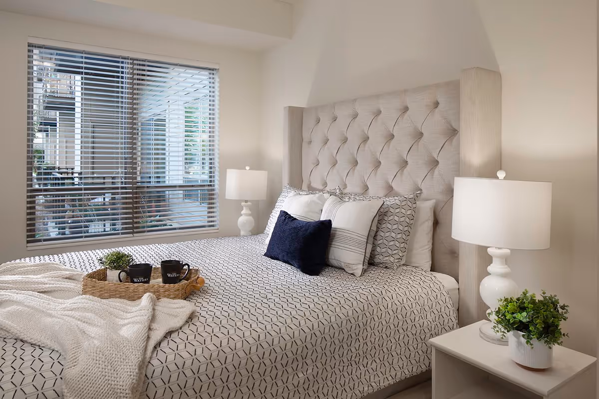 A neatly made bed with a tufted beige headboard in a bedroom. The bed has patterned bedding with several pillows, including a dark blue one. On the bed is a woven tray holding two black mugs and a small plant. There are two white bedside tables, each with a white lamp and a small green plant. A window with horizontal blinds is visible on the left side of the room.