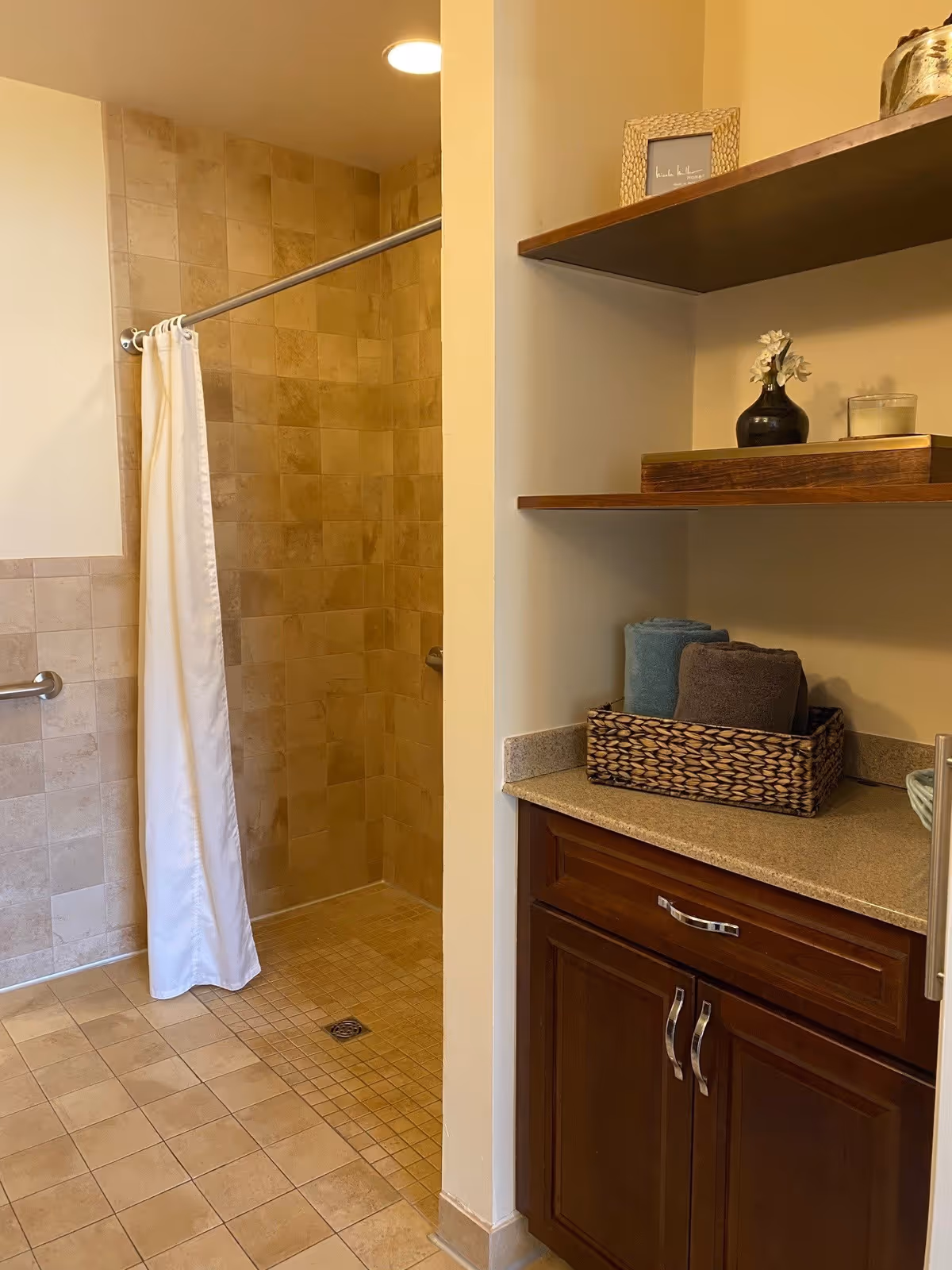 A bathroom with a tiled walk-in shower featuring a white shower curtain on a metal rod. To the right, there is a countertop with a woven basket holding rolled towels, a small vase with flowers, a candle, and a framed picture on a wooden shelf above the counter. The cabinetry below the counter is dark wood with silver handles.