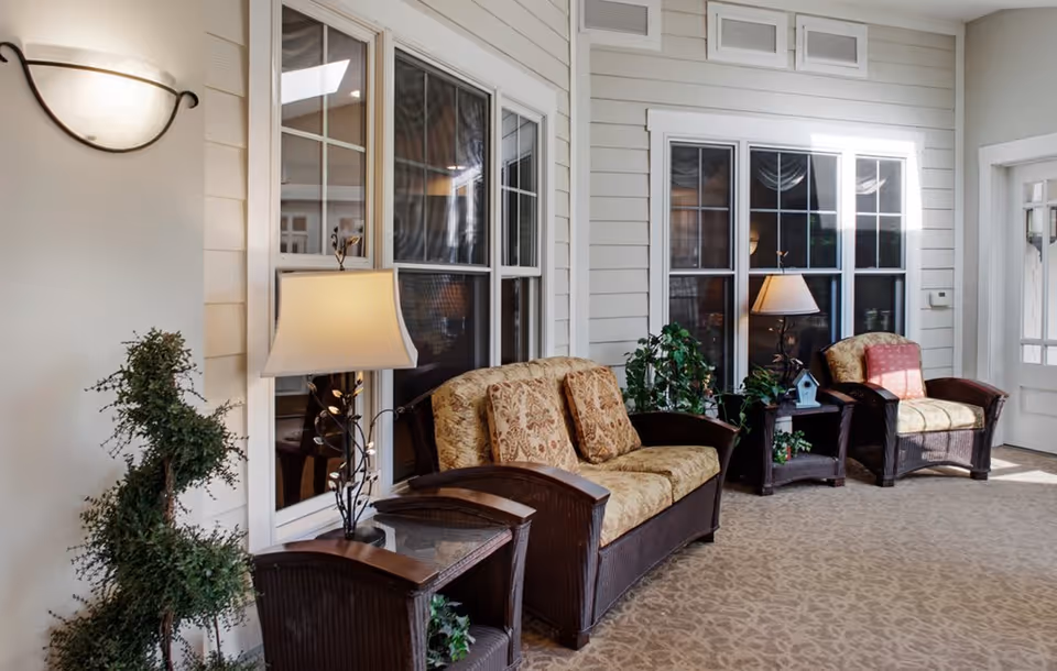 A cozy indoor seating area with two cushioned armchairs and a cushioned loveseat, all with wooden armrests and floral upholstery. There are two side tables with lamps and decorative plants placed near large windows with white frames. The walls are light-colored with horizontal paneling, and the floor is carpeted with a subtle pattern.