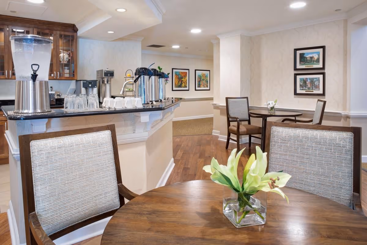Small dining/lounge area with round wooden tables and chairs, a beverage station with coffee and water dispensers, and framed artwork on the walls.
