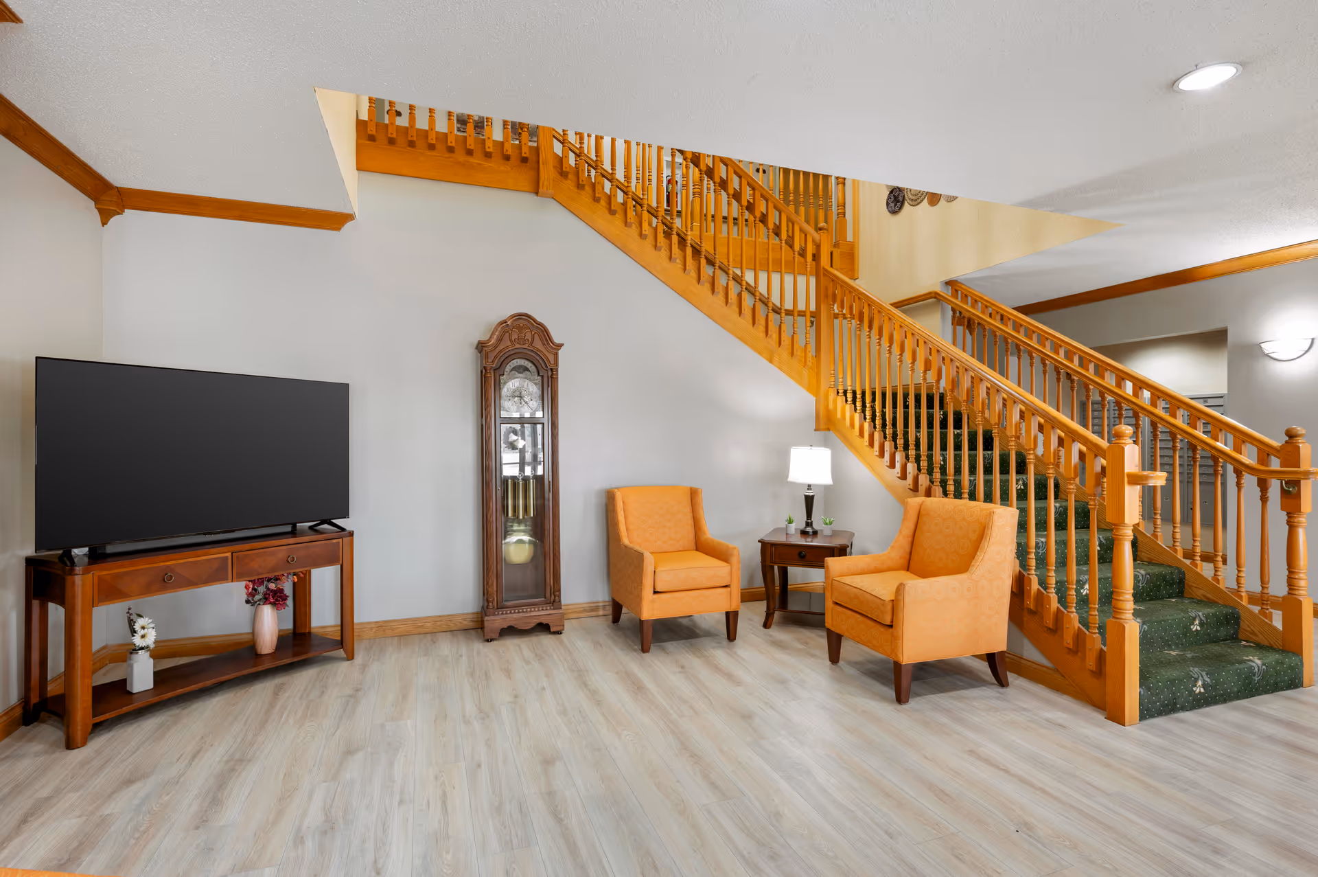A cozy living area featuring two orange upholstered armchairs with a small wooden side table and lamp between them, a wooden grandfather clock against the wall, a flat-screen TV on a wooden stand with decorative vases underneath, and a wooden staircase with green carpet leading to an upper floor.