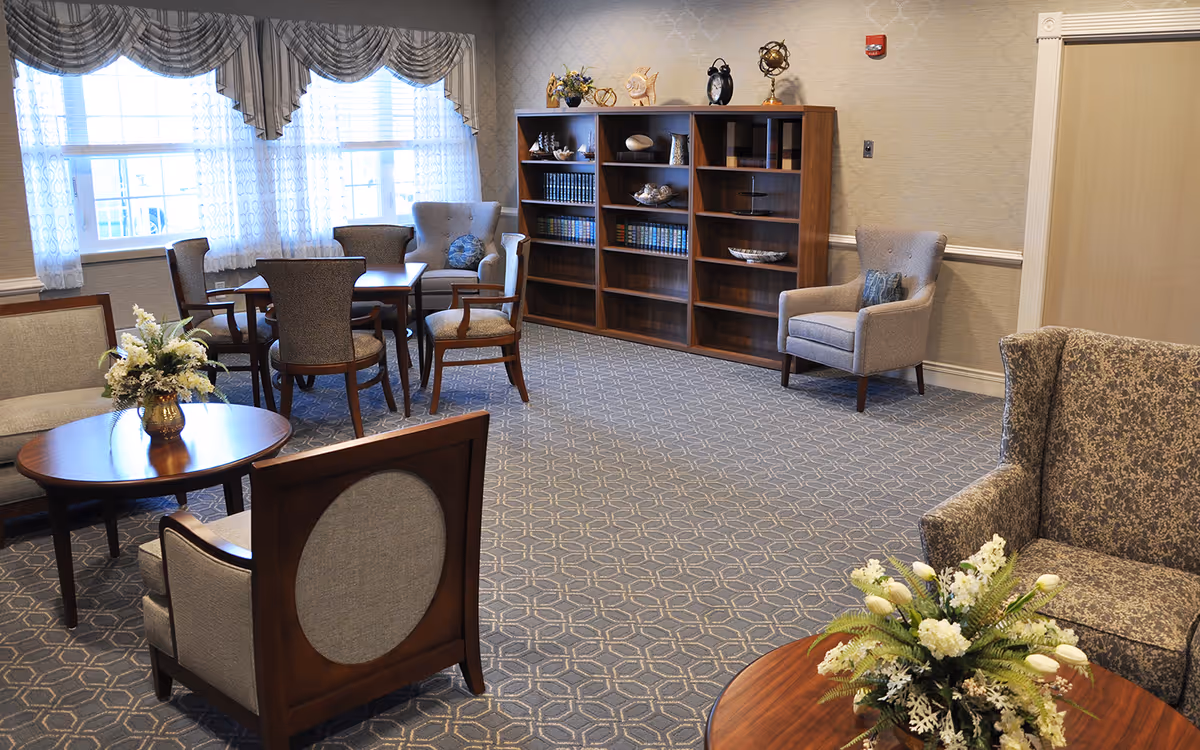 Well-lit senior living facility sitting room with armchairs, tables, a bookshelf, and floral centerpieces.