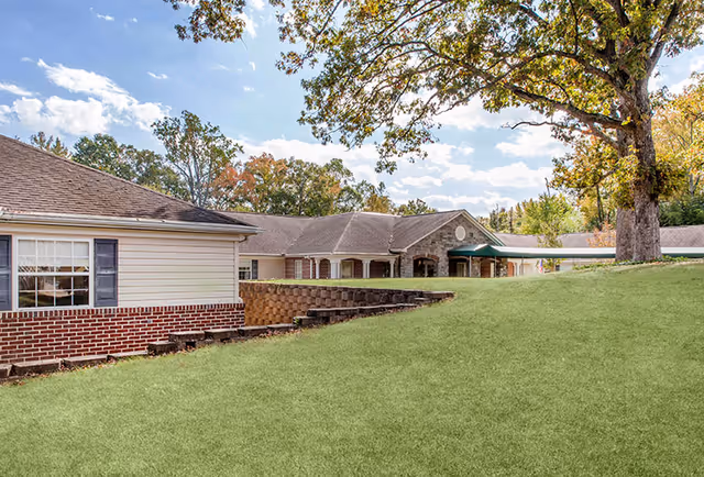 Exterior view of Charter Senior Living of Annapolis showing a single-story building with brick and siding, surrounded by green grass and large trees under a partly cloudy sky.