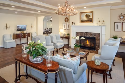 A cozy living room with light blue upholstered chairs and sofas arranged around a wooden coffee table and a fireplace with a lit fire. The room features a wooden side table with a plant, a chandelier with antler design, framed artwork above the fireplace, and soft lighting from wall sconces and lamps. The floor is wooden with a beige area rug, and an adjacent dining area is visible through an open doorway.
