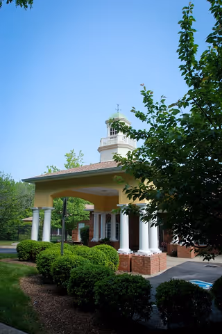Exterior view of a building entrance with white columns and a covered portico topped by a small cupola. The building is surrounded by green bushes and trees under a clear blue sky.