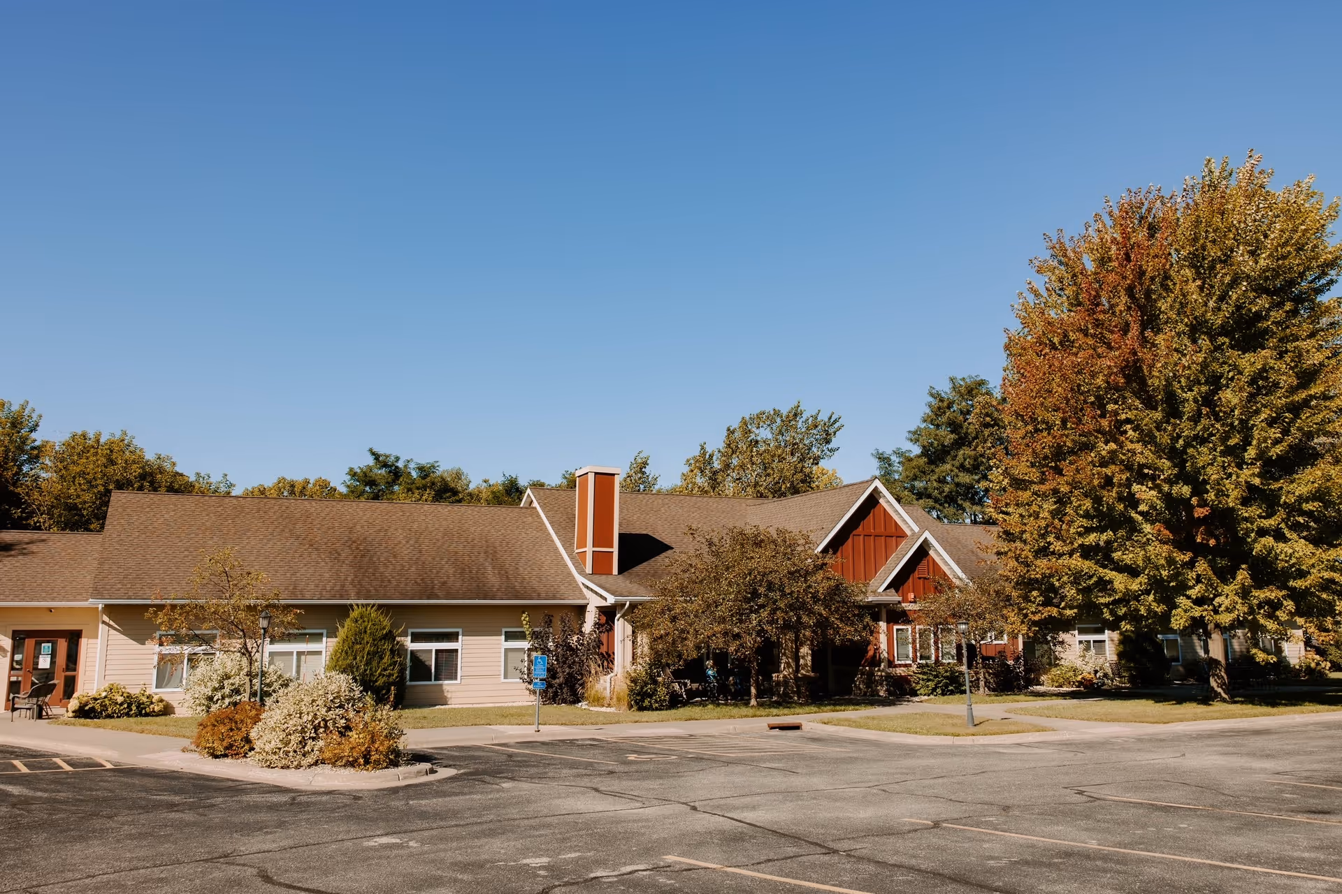 Exterior view of Valentines Assisted Living facility showing a single-story building with beige and red siding, surrounded by trees with autumn foliage and a parking lot in front under a clear blue sky.
