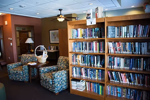 Interior view of a cozy common area with a wooden bookshelf filled with books, two patterned armchairs, a small round table with a basket on it, a floor lamp, and a ceiling fan with a light. The room has warm lighting and a framed picture on the wall.