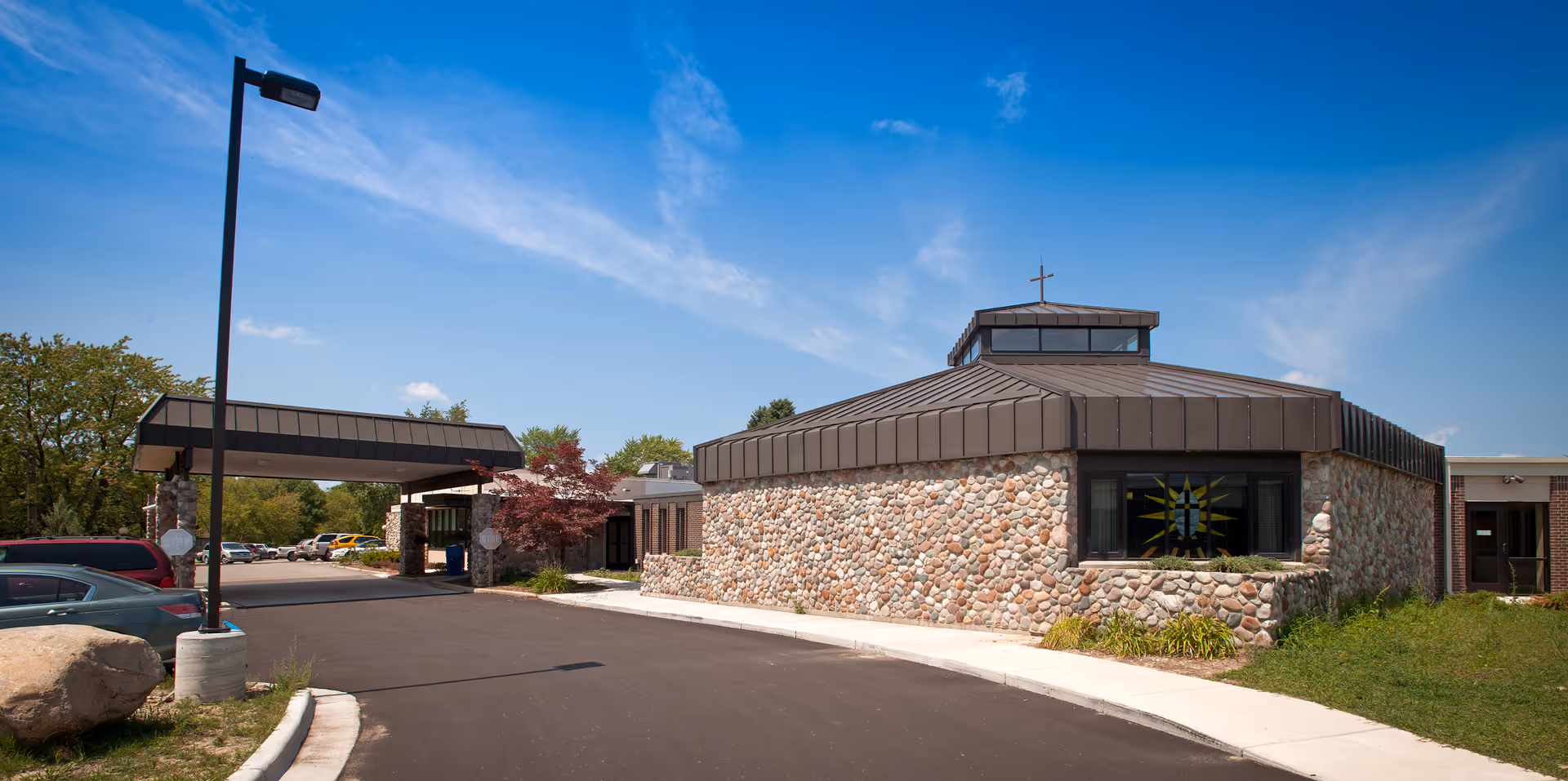 Exterior view of Wexford Senior Care Center showing a stone building with a brown metal roof and a cross on top, a covered entrance driveway, parked cars, and a clear blue sky.