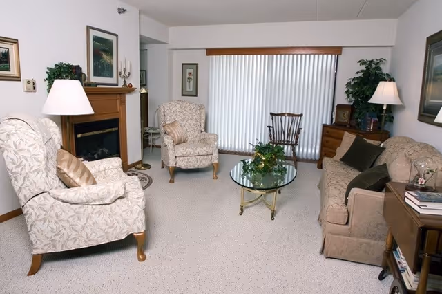 A cozy living room with two patterned armchairs, a beige sofa with dark cushions, a glass coffee table with a plant centerpiece, a wooden rocking chair, a fireplace with a wooden mantle, two table lamps, framed artwork on the walls, and vertical blinds covering a large window.