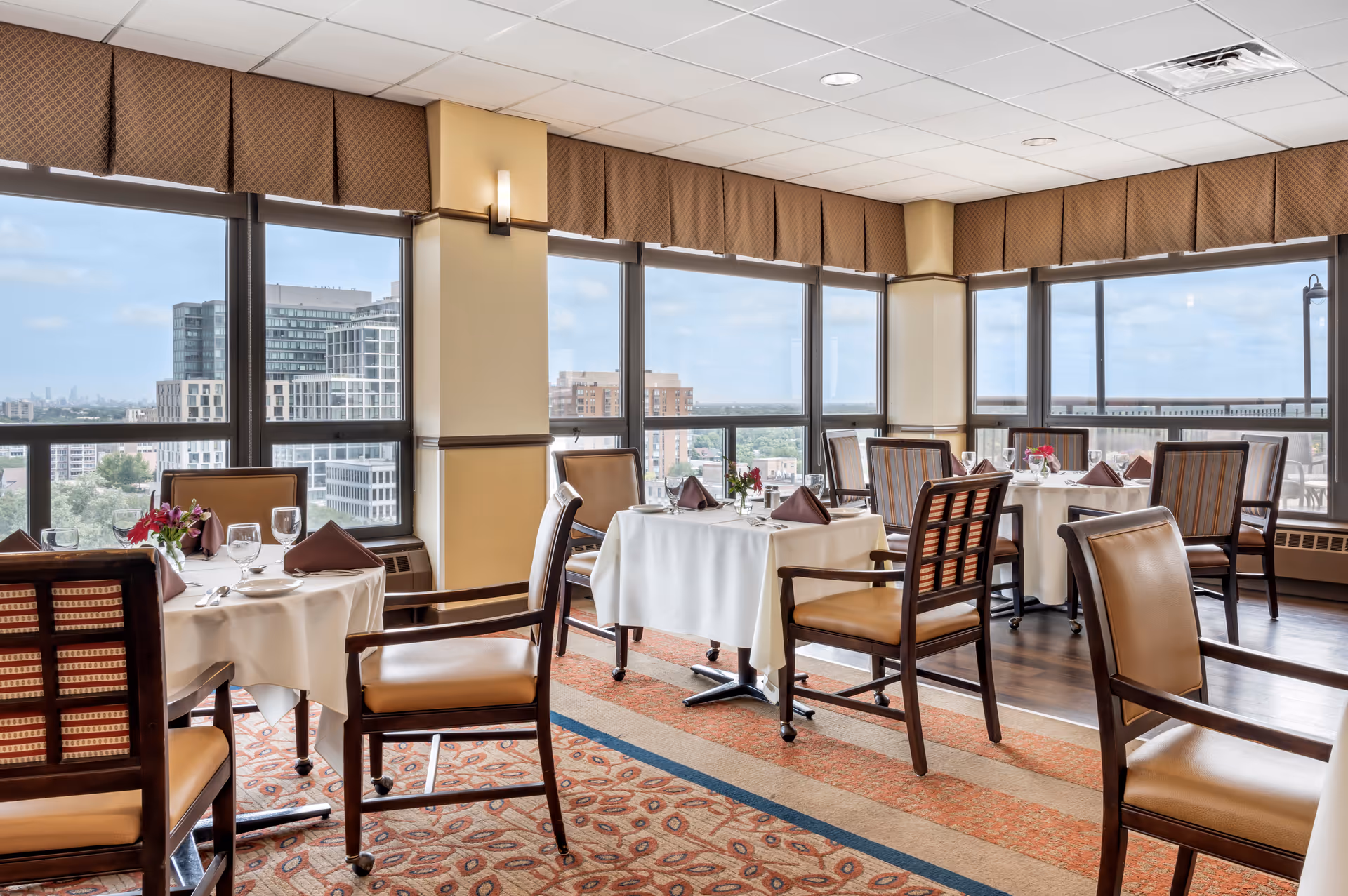 Bright dining room with set tables and chairs overlooking a cityscape through large windows.