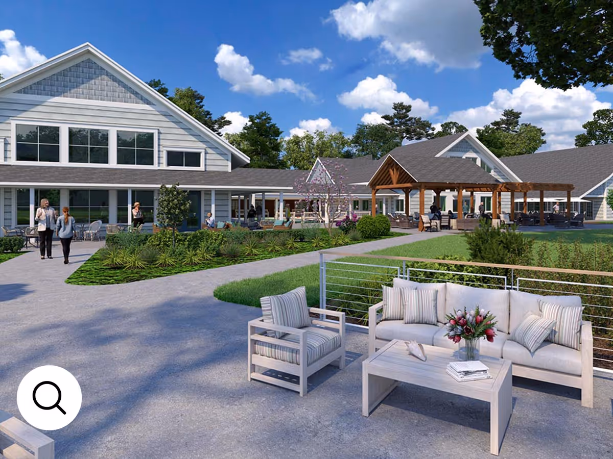 Outdoor seating area with cushioned chairs and a sofa around a coffee table with flowers, set on a paved patio in front of a large building with multiple windows and a wooden pergola. Several people are walking and sitting in the background, with green lawns, trees, and a blue sky with clouds.