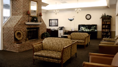 Carpeted lounge with a brick fireplace, patterned sofas and chairs, a fish tank, bookshelf and office equipment under soft ceiling lighting.