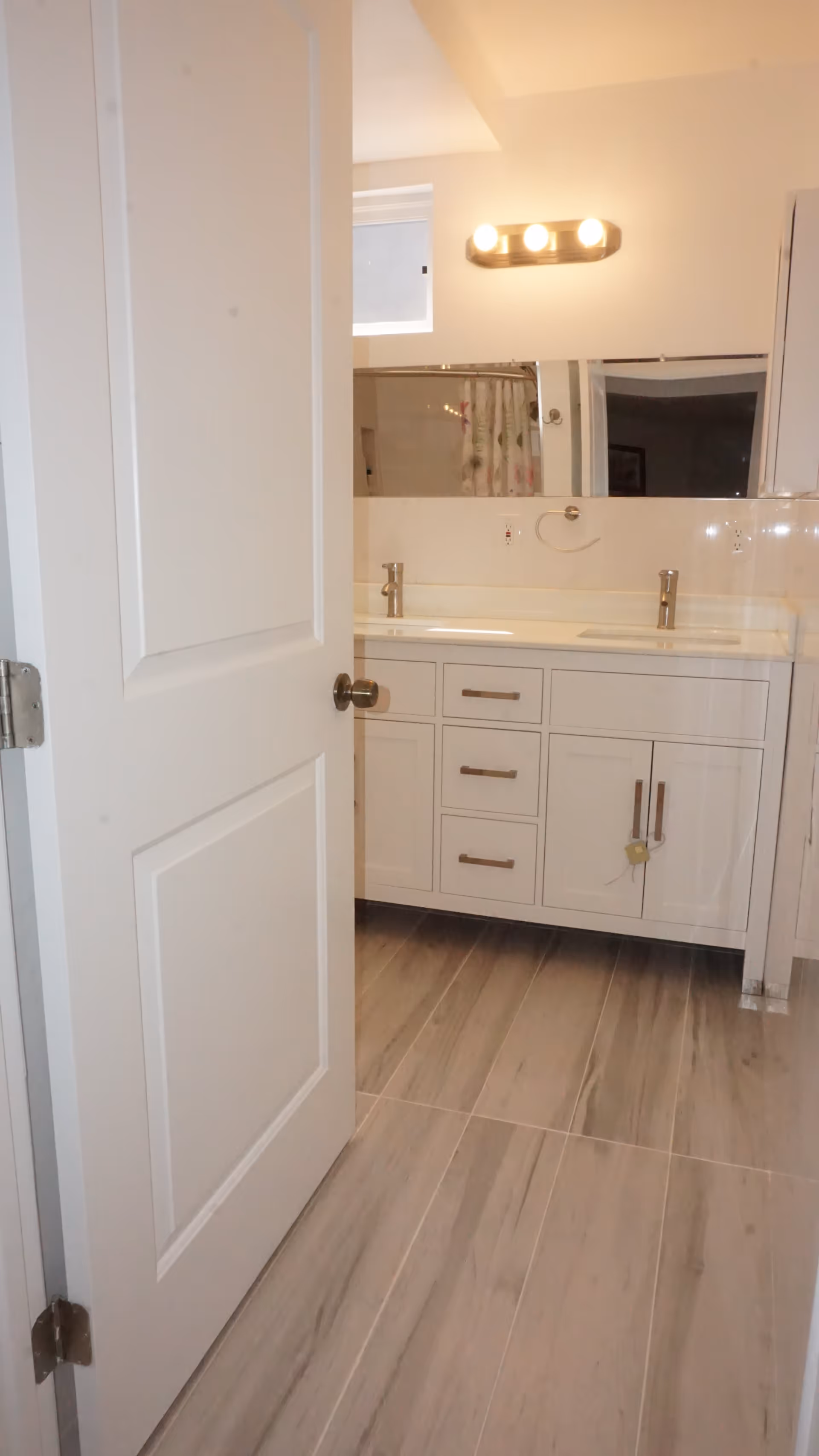 View into a bathroom with a partially open white door, showing a double sink vanity with white cabinets and silver handles, a large mirror above the sinks, a light fixture with three bulbs above the mirror, and a window near the ceiling. The floor has large gray tiles.