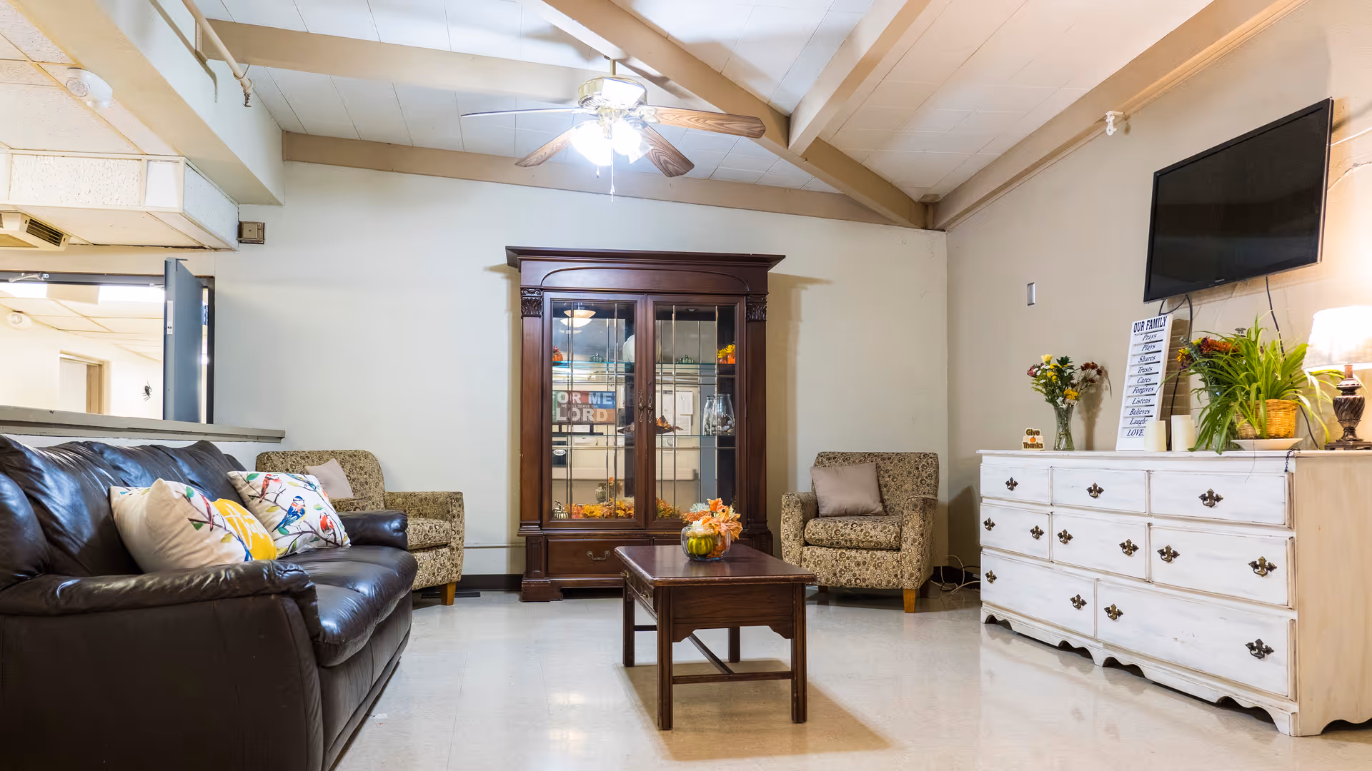 A cozy living room with a dark leather sofa adorned with colorful pillows, two patterned armchairs, a wooden coffee table with a small floral arrangement, a large wooden cabinet with glass doors, and a white dresser topped with plants, flowers, a lamp, and a wall-mounted flat-screen TV.