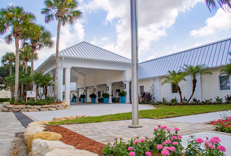 Exterior view of Buena Vida Estates featuring a covered entrance with white pillars, a metal roof, palm trees, landscaped garden beds with rocks and pink flowers, and a paved driveway.