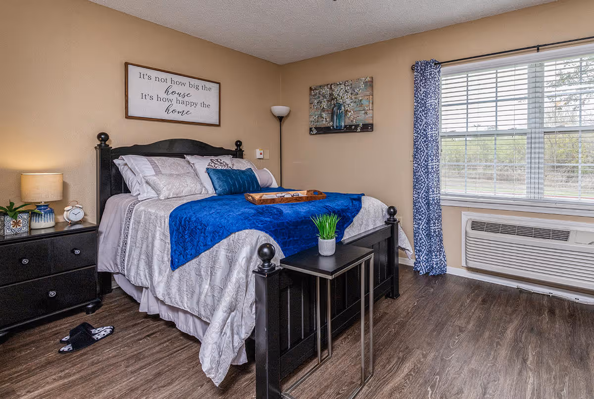 A cozy bedroom with a black wooden bed frame and a bed made with white and blue bedding. There is a small black side table with a green plant at the foot of the bed. To the left, a black nightstand holds a lamp, a small clock, and a plant. A framed sign above the bed reads, 'It's not how big the house, It's how happy the home.' A window with blue patterned curtains lets in natural light, and a floor lamp stands in the corner.