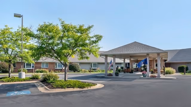 Exterior view of Brookdale Granger senior living facility showing a covered entrance with flags, surrounded by a parking area and landscaped greenery with trees and shrubs under a clear blue sky.
