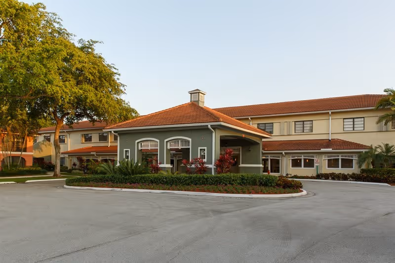 Exterior view of WillowBrooke Court Skilled Care facility showing a two-story building with a red-tiled roof, green and beige walls, surrounded by landscaped greenery and trees, with a circular driveway in front.