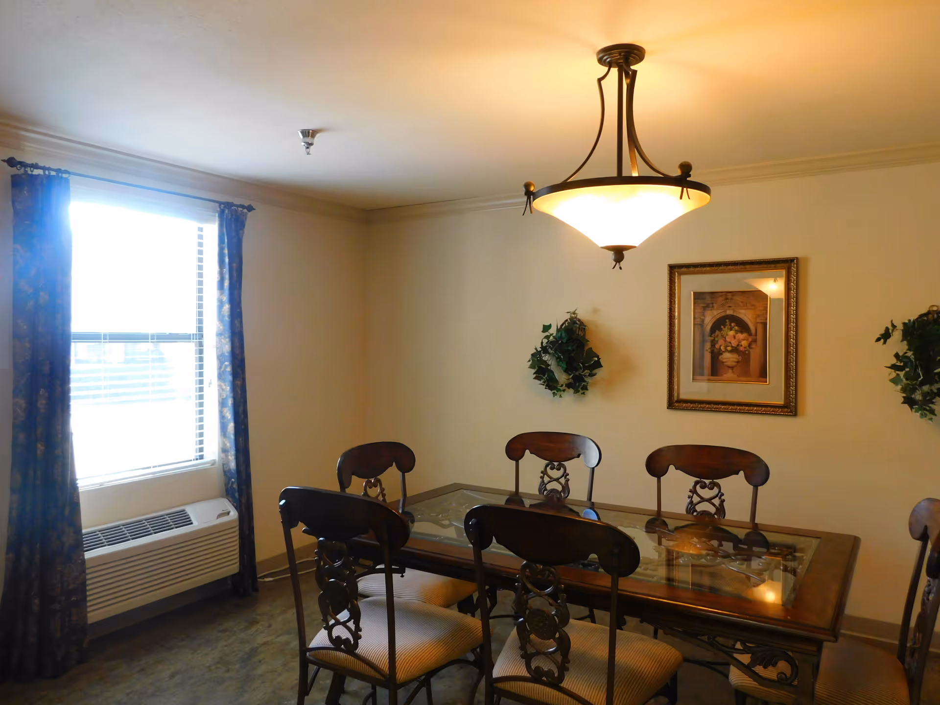 A dining room with a glass-top wooden table surrounded by six ornate wooden chairs with cushioned seats. The room has beige walls, a window with dark patterned curtains, an air conditioning unit below the window, a hanging ceiling light fixture, two small green wreaths on the wall, and a framed floral painting.