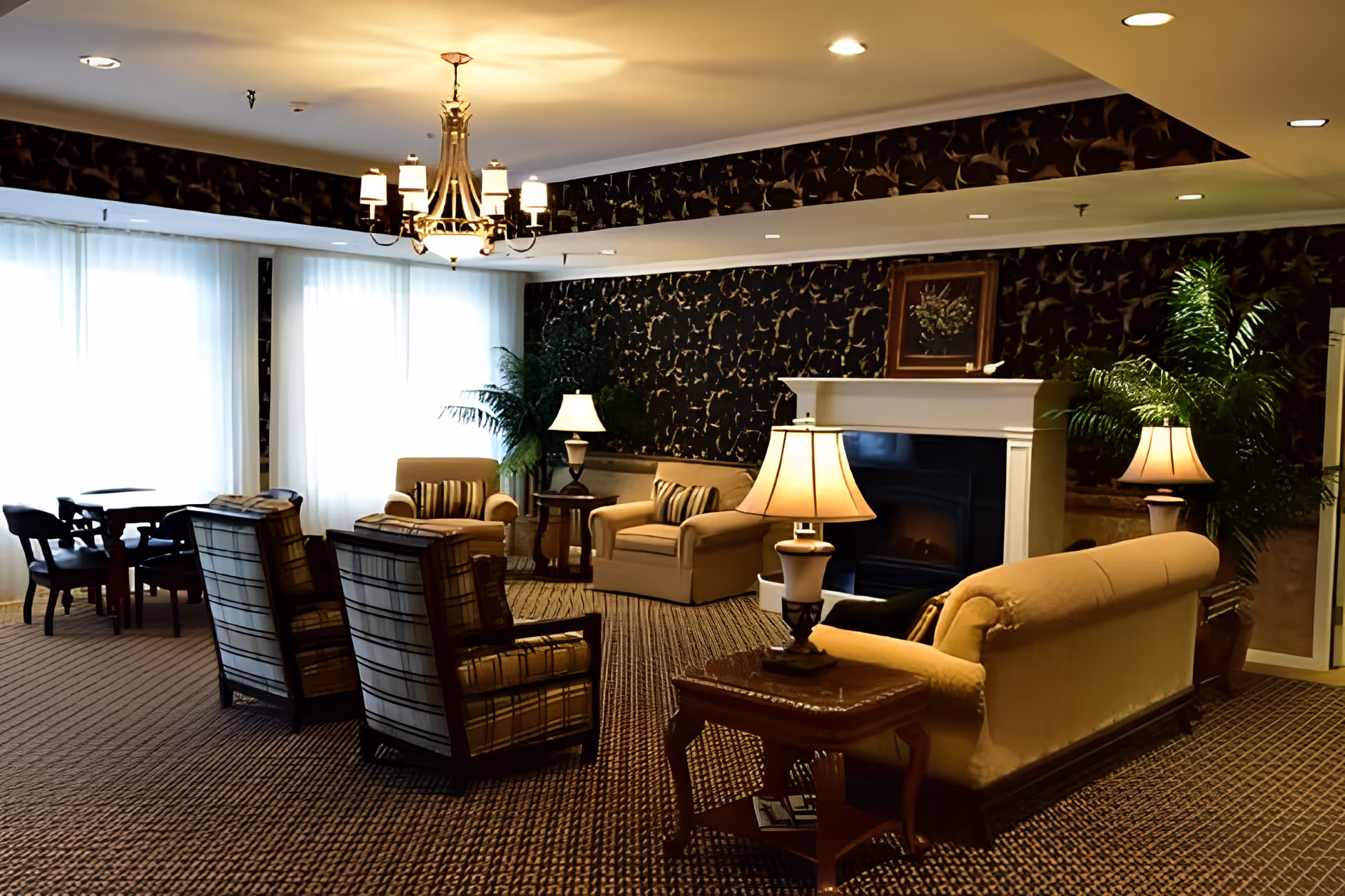 A cozy living room area with patterned carpet, multiple upholstered chairs and sofas, side tables with lamps, a fireplace with a framed picture above it, large windows with sheer white curtains, a chandelier, and potted plants.