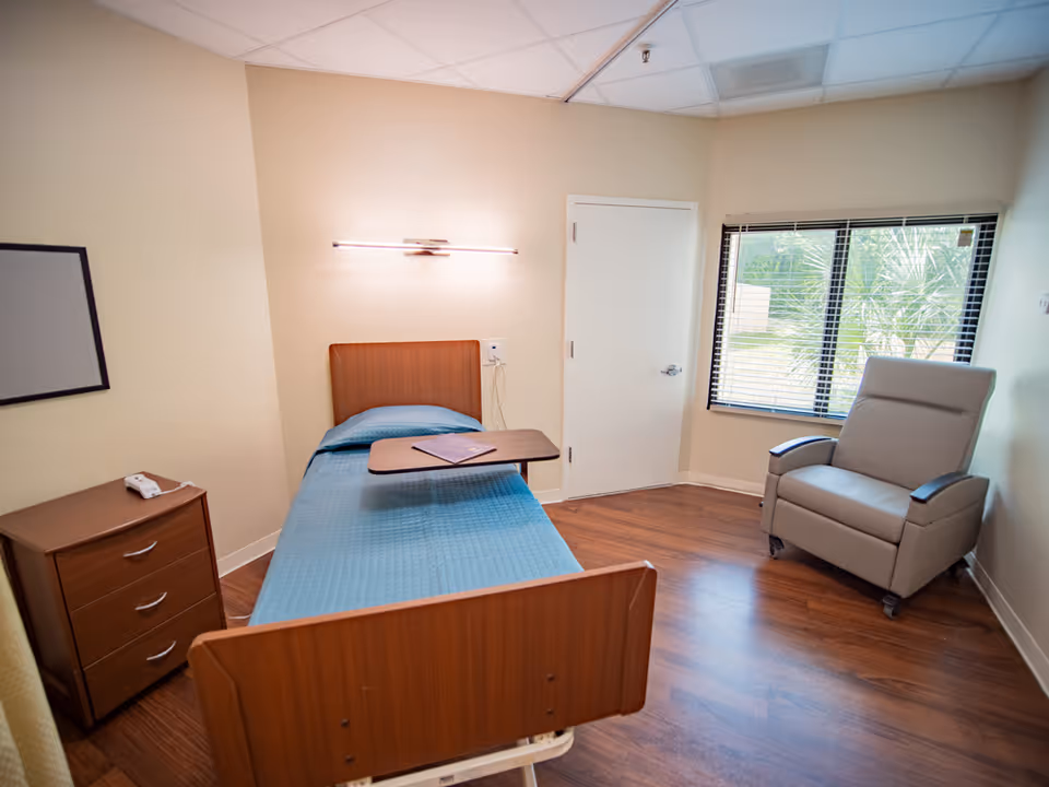 A simple, clean room in a healthcare facility with a single hospital bed covered with a blue blanket, a small wooden bedside table with a phone, a reclining chair near a window with blinds, and a door. The floor is wooden, and the walls are light-colored.
