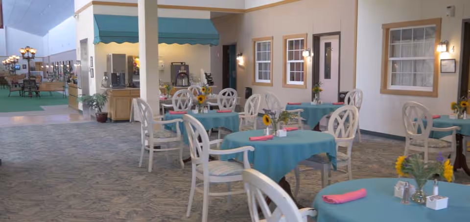 Interior view of a dining area in a senior living facility with round tables covered in teal tablecloths, each set with pink napkins and small flower arrangements featuring sunflowers. White chairs with patterned cushions surround the tables. The room has beige walls with windows and wall-mounted lights, and a beverage station is visible in the background.