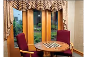 A cozy indoor seating area with a round wooden table featuring a chessboard on top, surrounded by two maroon upholstered chairs. The background shows three large windows with wooden frames and patterned valance curtains, offering a view of a green outdoor garden.
