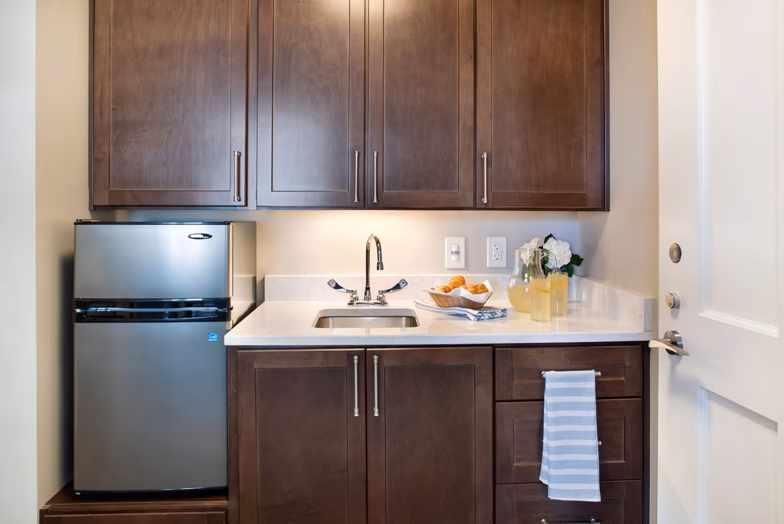 Small kitchen area with dark wooden cabinets, a stainless steel mini refrigerator, a white countertop with a sink, a pitcher of lemonade with two glasses, a basket of croissants, and a striped towel hanging on a drawer handle.