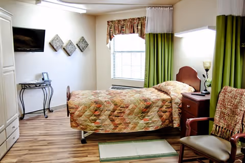 A single bed with a patterned bedspread in a nursing facility room. The room has wood flooring, a window with green and white curtains, a nightstand with a lamp and a flower vase, a chair with a blanket draped over it, a small table with a phone, and a wall-mounted TV.