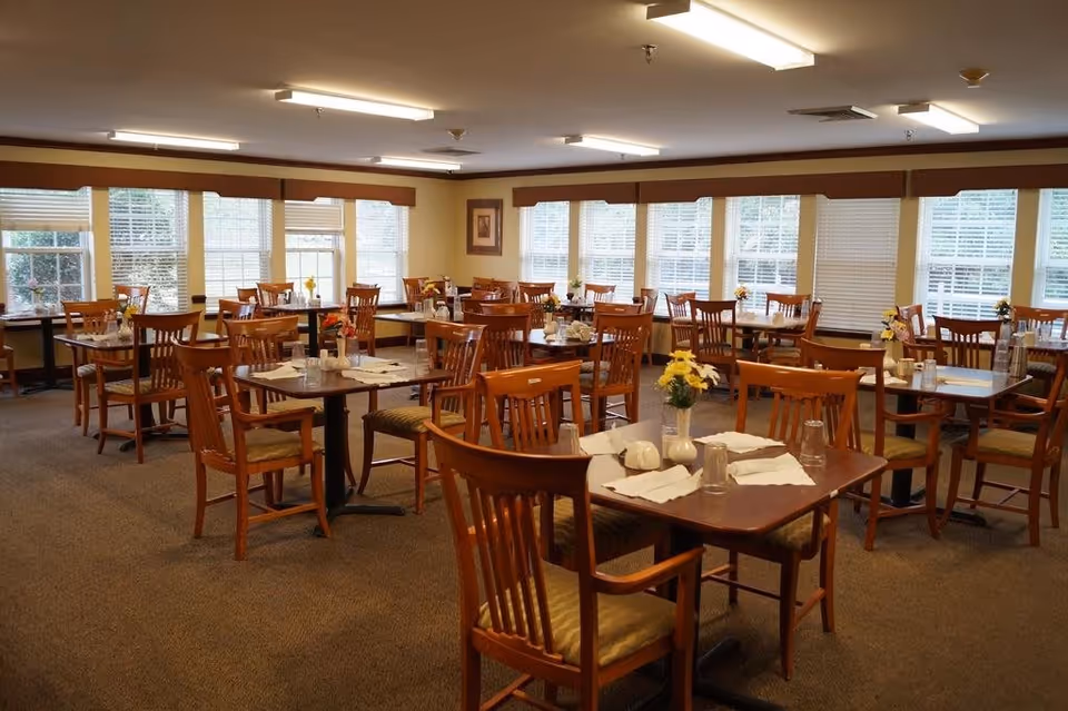 A dining room with multiple wooden tables and chairs arranged neatly. Each table is set with napkins, glasses, and small flower vases. Large windows with blinds allow natural light to fill the room.