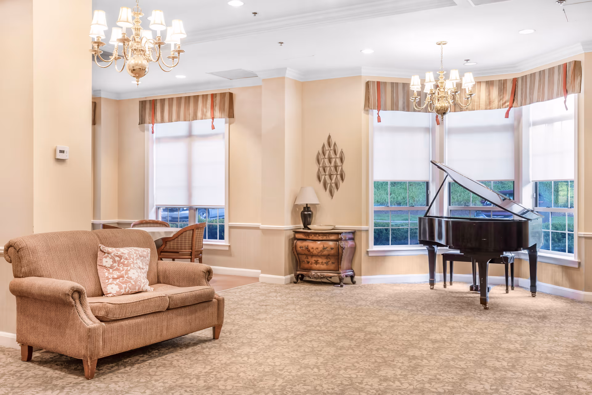 A bright and elegant living room area featuring a brown upholstered loveseat with a decorative pillow, a grand piano near large windows with white blinds and striped valances, a small wooden chest with a lamp and decorative wall art above it, and a chandelier hanging from the ceiling.