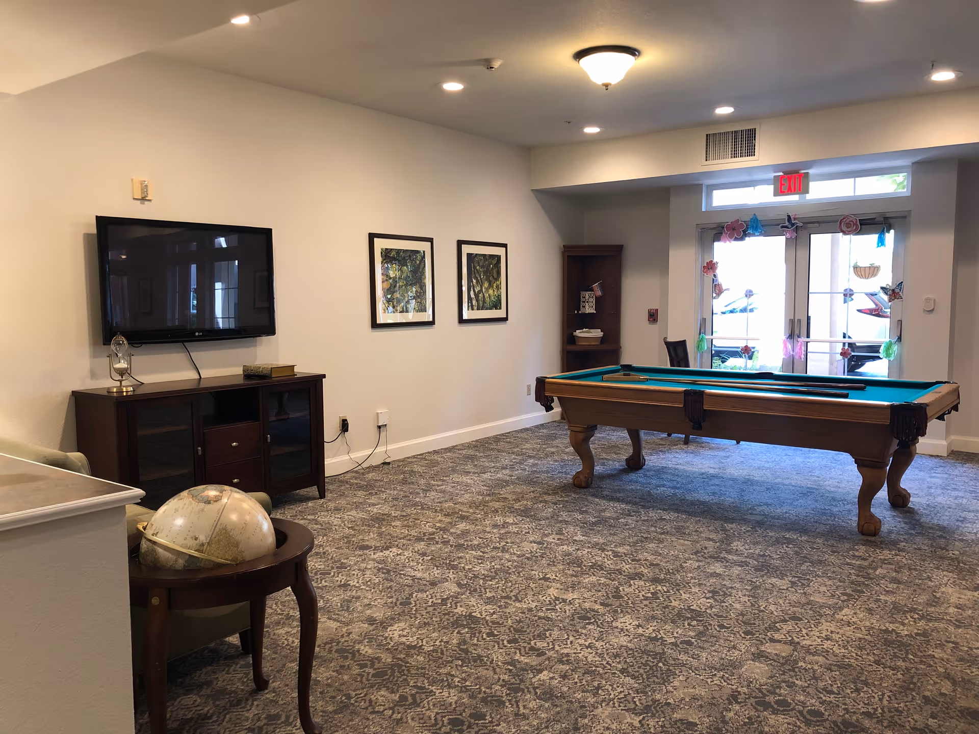 A recreational room with a pool table in the center, a wall-mounted flat-screen TV above a wooden cabinet, two framed pictures on the wall, a small round table with a globe on it, and glass double doors decorated with colorful paper flowers and butterflies.