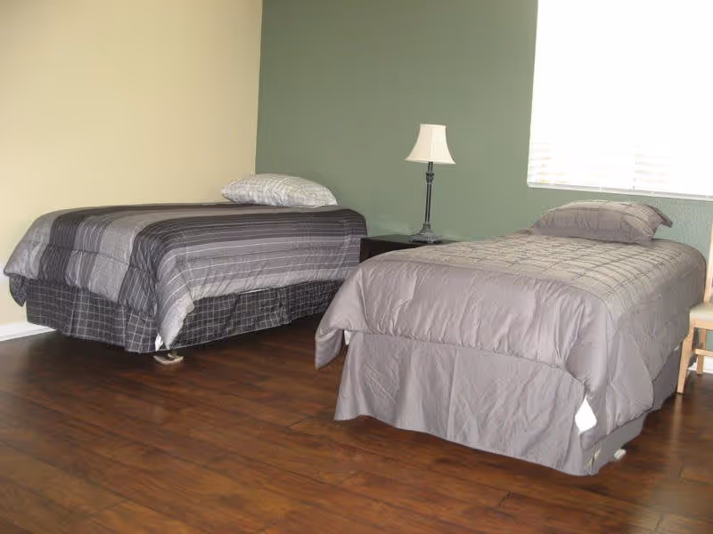 A simple bedroom with two single beds, each with gray bedding and pillows. There is a small nightstand between the beds with a lamp on it. The walls are painted in light yellow and green, and there is a window with blinds letting in natural light. The floor is wooden.