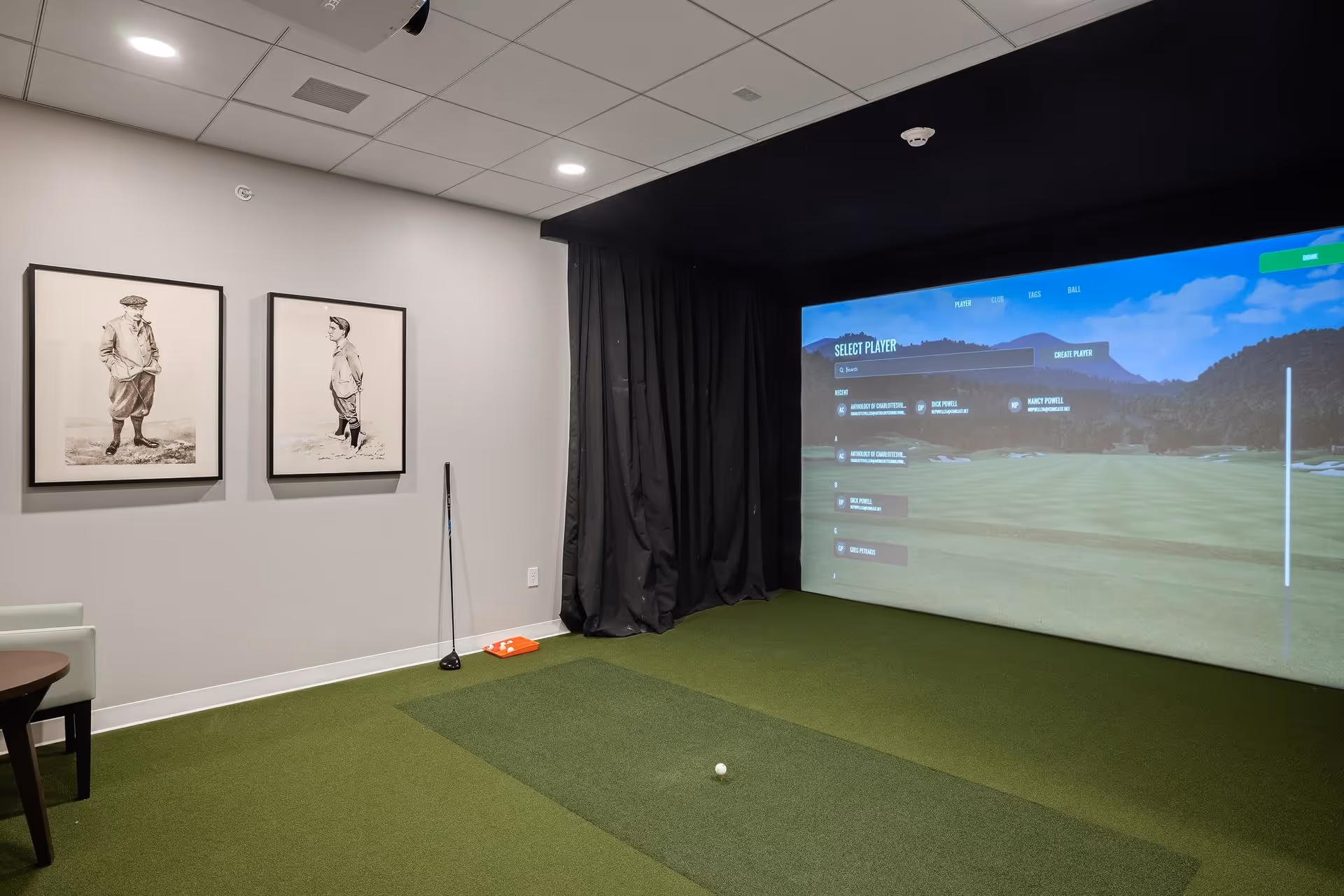 Indoor golf simulator room with green carpet flooring, a large screen displaying a golf course, two framed vintage golf player illustrations on the wall, a golf club leaning against the wall, and a small table with a chair.
