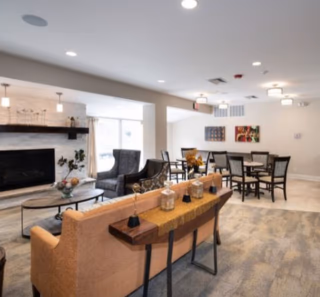 A bright and spacious senior living common area featuring a beige sofa with a wooden console table behind it, decorated with small vases and candles. In the background, there are several chairs and tables arranged for socializing or dining. A fireplace with a mantel and decorative items is visible on the left side, and the room is well-lit with ceiling lights.